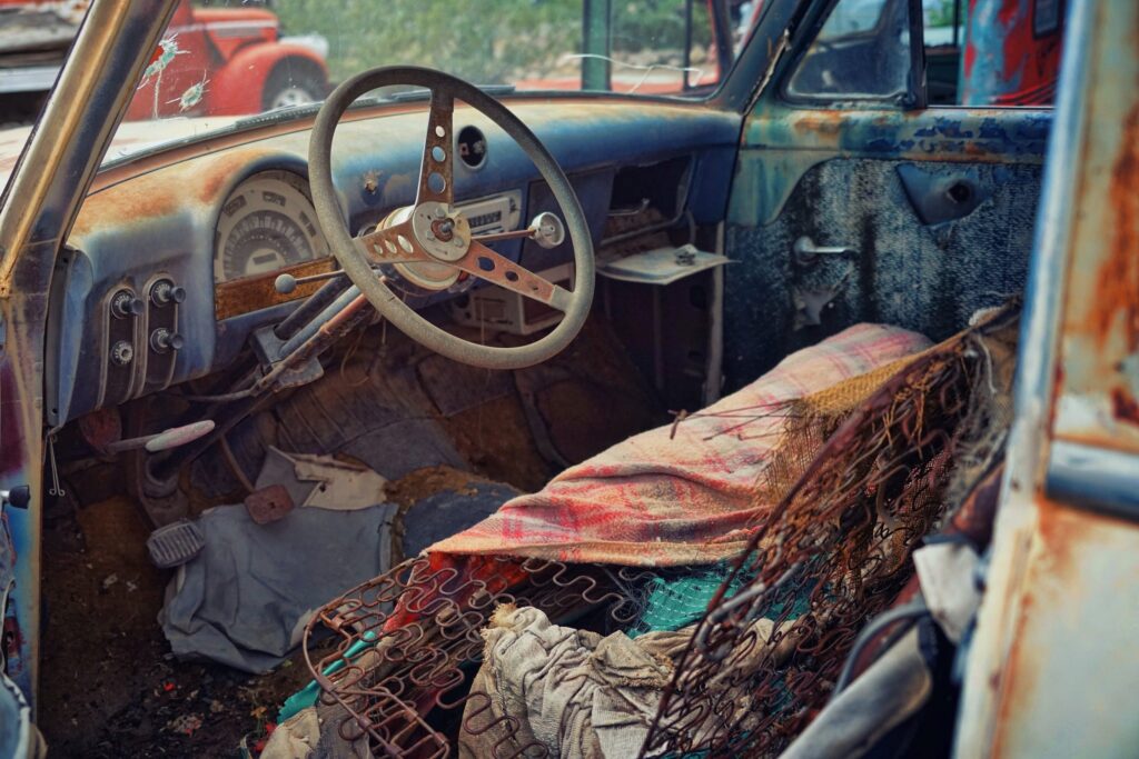 An interior shot of a junk car with a rusted steering wheel and seat.
