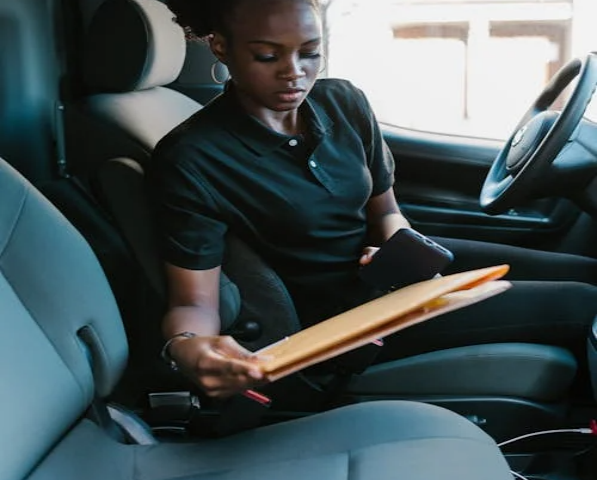 A woman sitting in a vehicle is holding a large envelope and a phone, appearing to handle delivery or paperwork tasks.