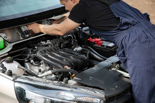 A mechanic in blue coveralls is working under the hood of a car, inspecting the engine components.