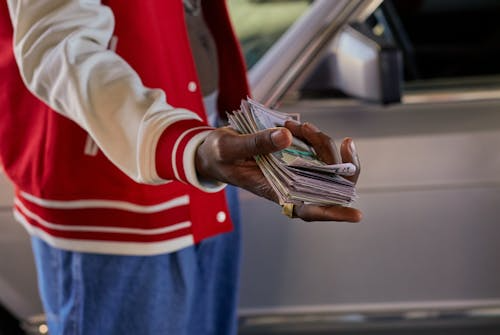 A person wearing a red jacket holds a large stack of cash beside a parked car.