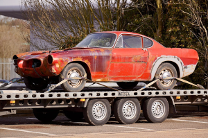 A rusty, vintage red car is strapped onto a flatbed trailer for transport.